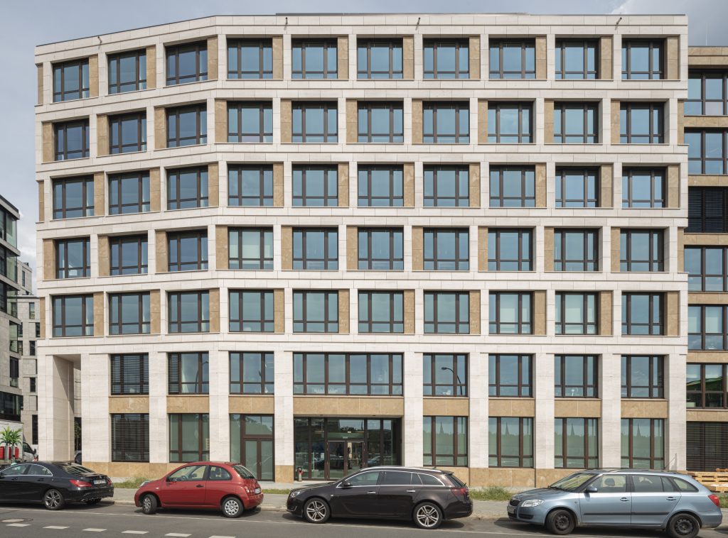 Ein symmetrisches, modernes Bürogebäude mit einer klaren Fassade aus weißen Steinrahmen, großen Glasfenstern und beigen Akzenten. Vor dem Gebäude stehen geparkte Autos entlang einer asphaltierten Straße.