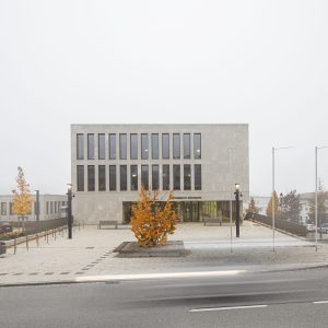 Modernes Gebäude mit heller Steinfassade und vertikalen Fensterreihen, umgeben von einem gepflasterten Vorplatz mit einem leuchtend orangefarbenen Baum in der Mitte.