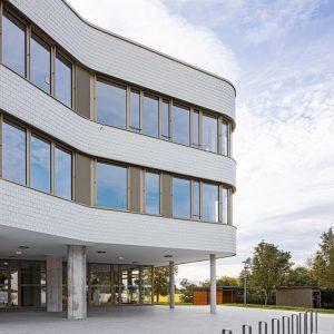 Modernes Gebäude mit geschwungener, weiß gekachelter Fassade und großen Fenstern. Der Eingangsbereich ist von schlanken Pfeilern gestützt, umgeben von Bäumen im Hintergrund.