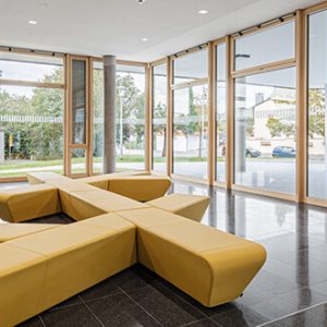 Moderner Innenraum mit bodentiefen Fenstern, die viel Tageslicht hereinlassen. Zentral steht eine gelbe, kreuzförmige Sitzbank auf glänzendem, dunklem Fliesenboden. Draußen sind Bäume und ein urbanes Umfeld sichtbar.