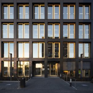 Frontansicht eines modernen Bürogebäudes mit einer dunklen Fassade, die von rechteckigen, bodentiefen Fenstern durchbrochen wird. Die Fenster reflektieren das warme Licht der tief stehenden Sonne, während ein gepflasterter Eingangsbereich mit Rampen und dezenten Beleuchtungen den Zugang markiert.