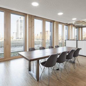 Moderner Besprechungsraum mit einem großen Holztisch und acht gepolsterten Stühlen. Große Holzrahmenfenster lassen viel Tageslicht herein und bieten Blick auf umliegende Gebäude und eine Grünfläche.