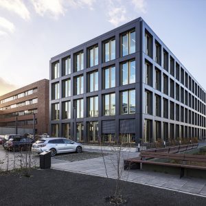 Ein modernes Bürogebäude mit dunkler Fassade und großen Fenstern mit goldenen Rahmen, aufgenommen bei Sonnenuntergang, wodurch ein warmes Licht die Umgebung beleuchtet. Im Vordergrund befinden sich ein Parkplatz mit mehreren Autos, eine Bank auf einem gepflasterten Weg und spärlich bepflanzte Grünflächen.