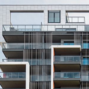 Detailansicht der oberen Etagen eines modernen Wohngebäudes mit großzügigen Balkonen, bodentiefen Fenstern und auffälligen vertikalen Metallstreben als Gestaltungselemente. Die Dachpartie umfasst offene Rahmenstrukturen und Glasgeländer.