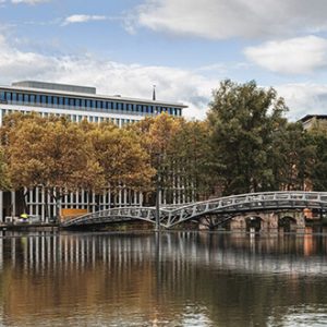 Modernes Bürogebäude mit großen Glasfassaden, umgeben von herbstlich gefärbten Bäumen. Im Vordergrund ein ruhiger Fluss, über den eine geschwungene Metallbrücke führt.