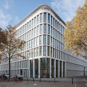 Moderne Bürogebäude-Fassade mit abgerundeten Ecken und vertikalen Fensterstreben, umgeben von herbstlichen Bäumen. Im Vordergrund sind Fahrräder und Pflastersteine sichtbar.
