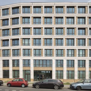 Ein symmetrisches, modernes Bürogebäude mit einer klaren Fassade aus weißen Steinrahmen, großen Glasfenstern und beigen Akzenten. Vor dem Gebäude stehen geparkte Autos entlang einer asphaltierten Straße.