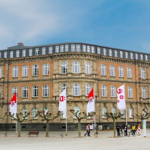 Historisches Gebäude mit roter Klinkerfassade und großen weißen Fenstern, verziert mit klassischen Steinelementen. Davor stehen kahle, symmetrisch angeordnete Bäume und mehrere Fahnen mit roten und weißen Logos.