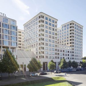 Moderner Hotelkomplex mit einer Fassade aus hellem Putz und großen, symmetrisch angeordneten Fenstern. Die Baukörper mit abgerundeten Ecken sind terrassenartig angeordnet und von einer begrünten Umgebung mit Bäumen und parkenden Autos umgeben.