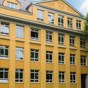 Gelbes mehrstöckiges Gebäude mit klassizistischen Elementen, symmetrischer Fassade und weißen Fensterrahmen umgeben von grünen Bäumen. Giebel und Dachfenster verleihen dem Bau ein charakteristisches Erscheinungsbild.
