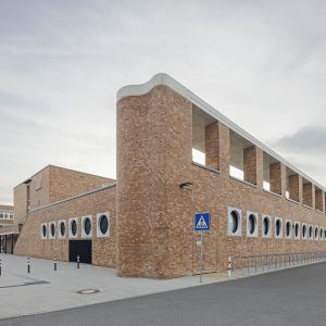 Ein langes Gebäude mit einer großen Backsteinfassade und runden Fenstern. Davor befinden sich ein Verkehrsschild für einen Fußgängerüberweg und zahlreiche Fahrradständer.