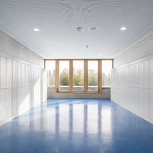 Graue Schließfächer in einem Schulflur mit blauem Boden und Holzfenstern am Ende des Gangs.