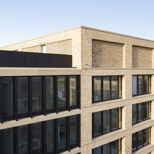 Moderne Gebäude mit hellen Klinkerziegel-Fassaden und großen, schwarzen Fenstern, ergänzt durch dekorative, gemusterte Ziegelgitter in den oberen Etagen unter blauem Himmel.