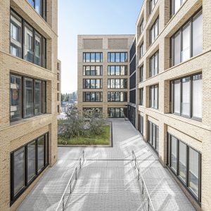 Innenhof eines modernen Bürokomplexes mit Ziegelfassaden und großen schwarzen Fensterrahmen. Der Hof ist mit hellgrauem Pflaster gestaltet, enthält ein begrüntes Beet mit kleinen Bäumen und Grasflächen.