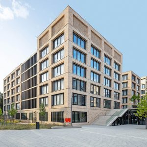 Zeitgemäßes Bürogebäude mit heller Klinkerfassade und großen, symmetrisch angeordneten Glasfenstern, die viel Licht ins Innere lassen. Im Vordergrund befinden sich ein gepflasterter Platz, ein breiter Treppenaufgang sowie Grünflächen mit jungen Bäumen, die das moderne Design ergänzen.