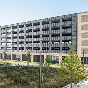 Moderne Architektur mit einem mehrstöckigen Parkhaus aus dunklen Fassadenelementen und seitlichen Backsteinflächen. Im Vordergrund befinden sich begrünte Außenanlagen mit jungen Bäumen und gepflasterten Wegen.