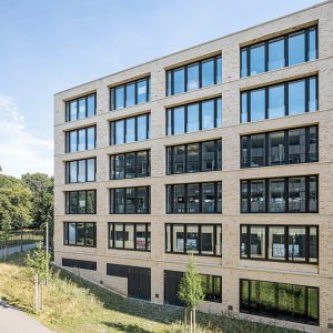 Moderne Gebäudefassade aus hellen Ziegelsteinen mit großen, symmetrisch angeordneten Fenstern und klarer, geradliniger Architektur. Vor dem Gebäude verläuft ein begrünter Weg mit jungen Bäumen und gepflegter Vegetation.
